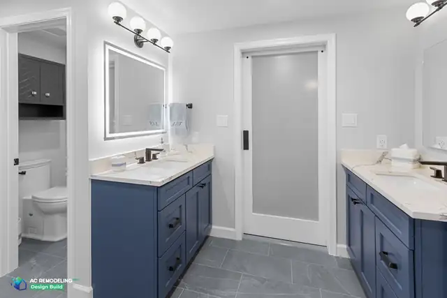 Twin navy vanities with quartz counters, globe sconces and backlit mirrors in a Gaithersburg, MD primary bath